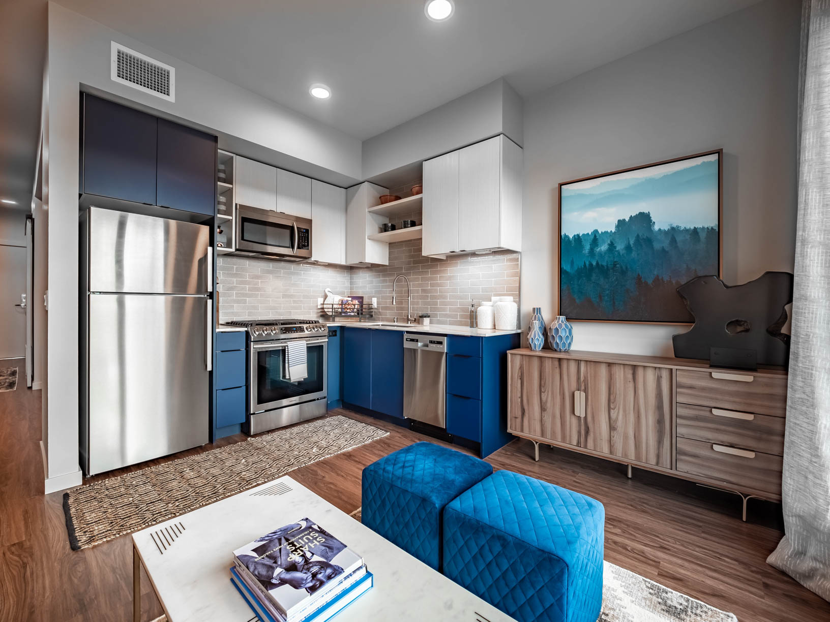 Furnished L-shaped apartment kitchen and living area with wood flooring, stainless steel appliances, two-tone cabinets, and tile backsplash.