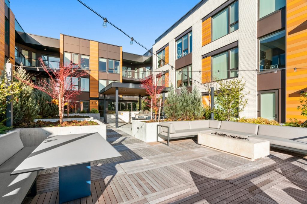 Outdoor courtyard with covered grill area, sectional couches, dining tables, a fire pit, raised planters, and string lighting.