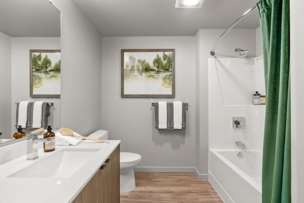 Apartment bathroom with wood flooring, an under-mounted sink, large wall mirror, and a shower and bathtub combo with curved shower rod.